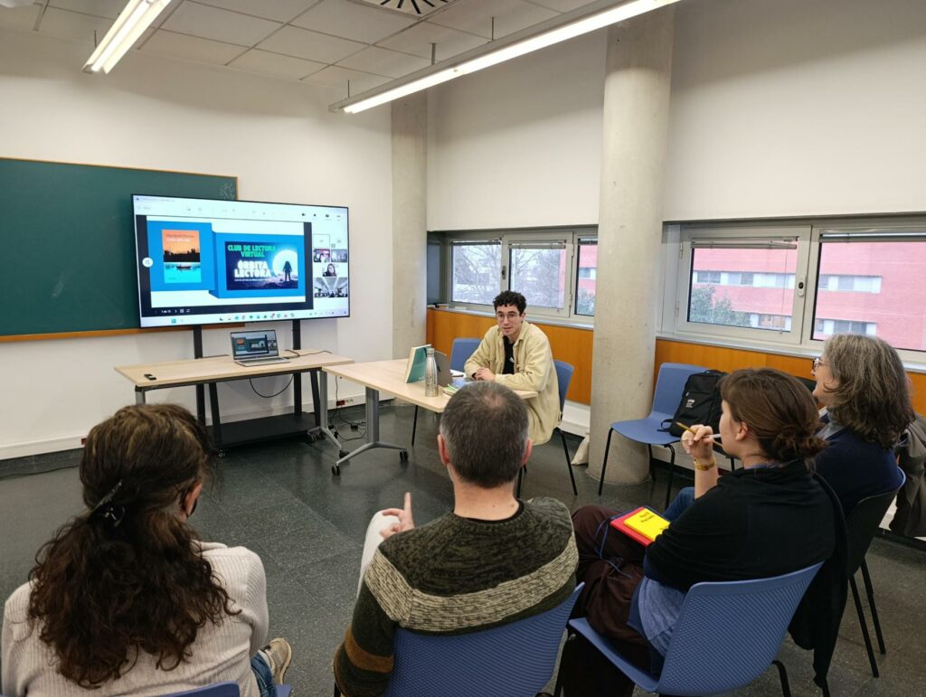 Una trobada presencial i virtual del club de lectura «Òrbita lectora» en una aula, amb un grup de persones assegudes en semicercle escoltant un jove al fons.