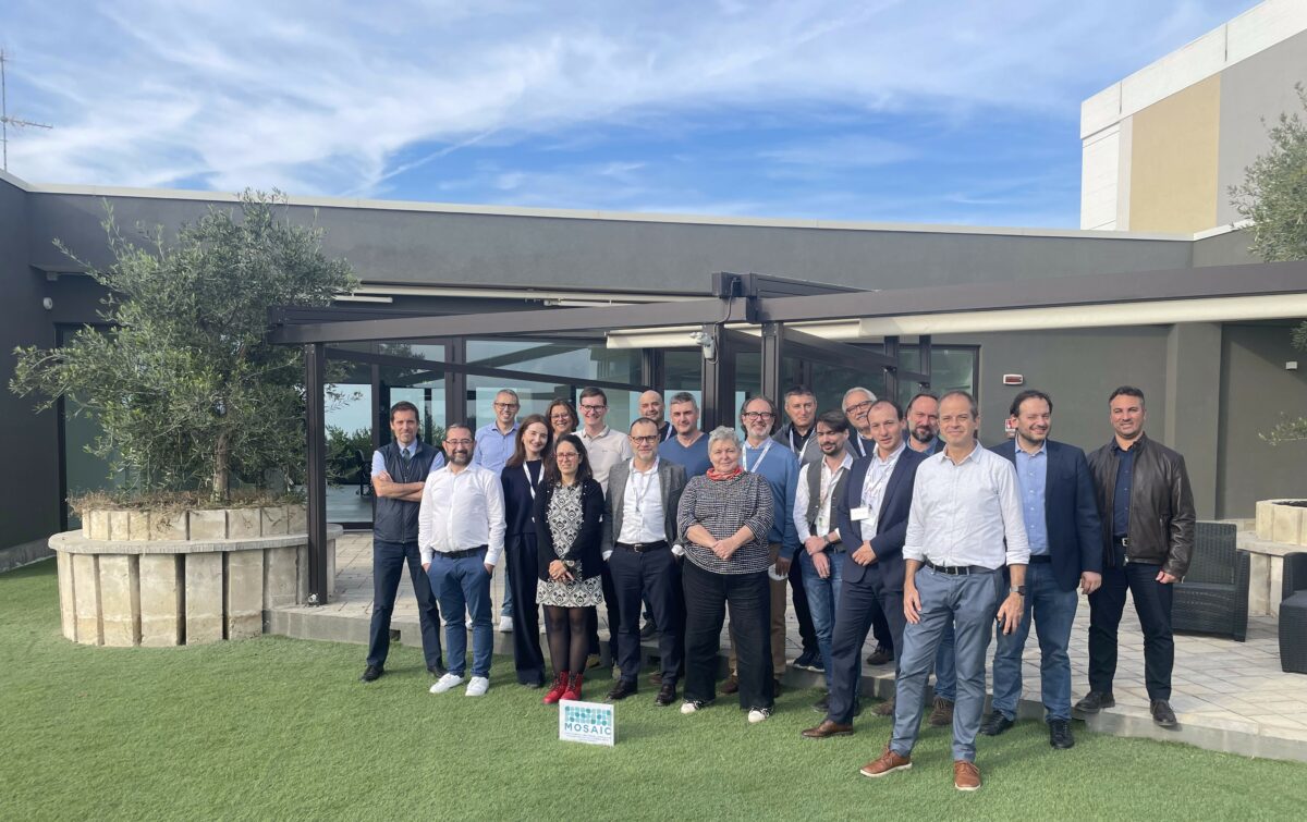 A group photo of the MOSAIC project team, consisting of around 16 people, standing outdoors on a green lawn in front of a modern building with large glass windows and a clear blue sky above. The team members are dressed in professional, business-casual attire, smiling at the camera, and some are wearing identification lanyards. A small sign with the MOSAIC project logo is displayed in front of the group on the grass