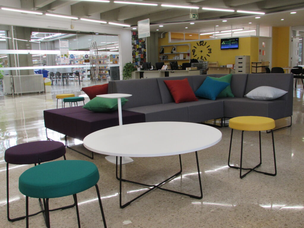 Fotografia del vestíbul de la Biblioteca de Ciència i Tecnologia de la UAB. En primer pla es veu una taula rodona blanca amb tres tamborets, i darrera hi ha un sofà gris amb coixins de colors llisos. Més al fons es veu el taulell de préstec i la sala de silenci, separada per una paret de vidre.
